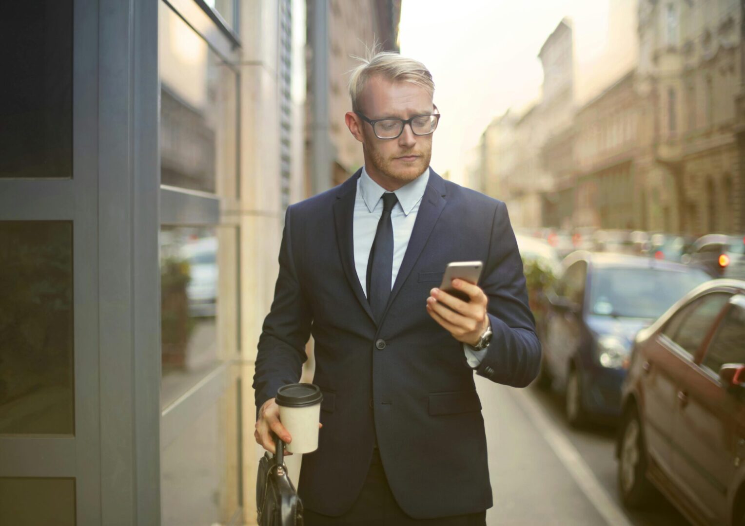 A man in suit and tie holding a coffee cup while looking at his phone.