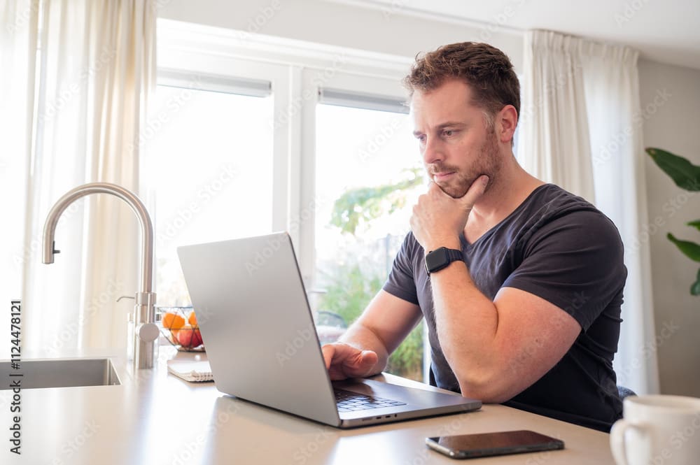 Man thoughtfully working on a laptop at home with a cup of coffee nearby.