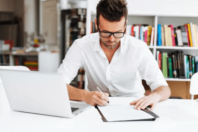 Man in white shirt writing on clipboard at desk with laptop.