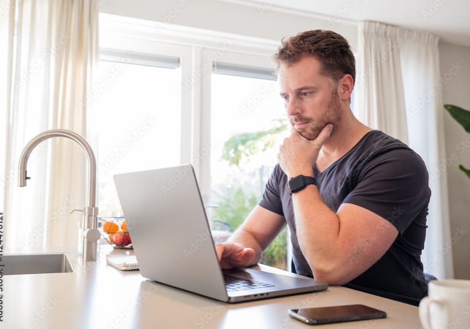Man thoughtfully working on a laptop at home with a cup of coffee nearby.