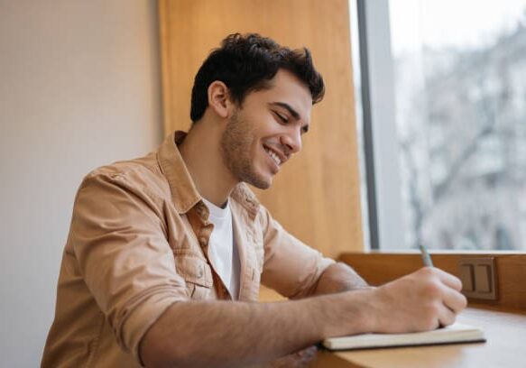 Happy Indian business man writing on notebook, working project, planning strategy in office. Smiling university student studying, exam preparation, taking notes. Education concept